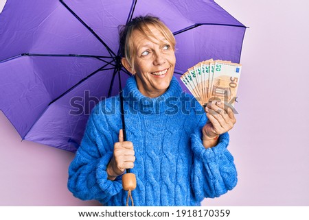 Middle age blonde woman holding umbrella and 50 euros banknotes smiling looking to the side and staring away thinking. 