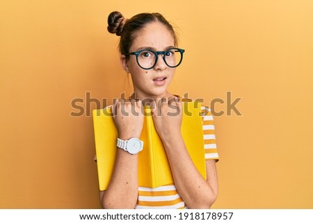Beautiful brunette little girl wearing glasses and holding book clueless and confused expression. doubt concept. 