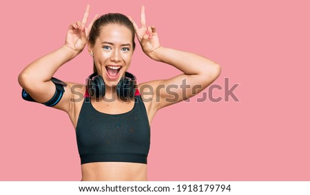 Beautiful blonde woman wearing gym clothes and using headphones posing funny and crazy with fingers on head as bunny ears, smiling cheerful 
