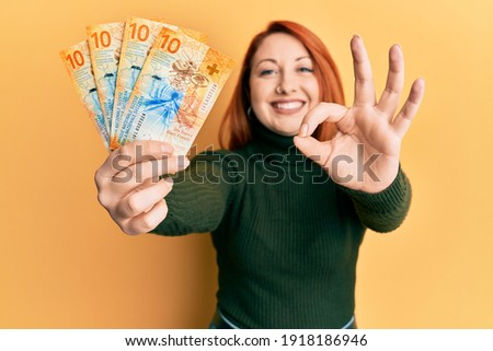 Beautiful redhead woman holding 10 swiss franc banknotes doing ok sign with fingers, smiling friendly gesturing excellent symbol 