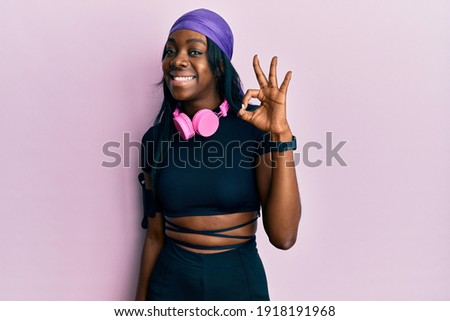 Young african american woman wearing gym clothes and using headphones smiling positive doing ok sign with hand and fingers. successful expression. 
