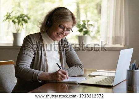 Smiling mature woman wearing headphones writing notes, using laptop, sitting at table at home, aged female wearing glasses listening to lecture, watching webinar, studying online, learning language Royalty-Free Stock Photo #1918212572
