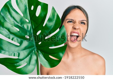 Young brunette woman holding green plant leaf close to beautiful face angry and mad screaming frustrated and furious, shouting with anger looking up. 
