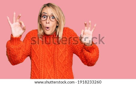Beautiful blonde woman wearing casual clothes and glasses looking surprised and shocked doing ok approval symbol with fingers. crazy expression 