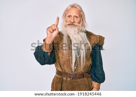 Old senior man with grey hair and long beard wearing viking traditional costume pointing finger up with successful idea. exited and happy. number one. 