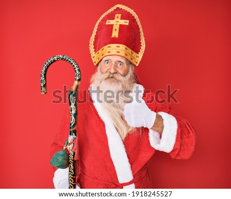 Old senior man with grey hair and long beard wearing traditional saint nicholas costume doing happy thumbs up gesture with hand. approving expression looking at the camera showing success. 