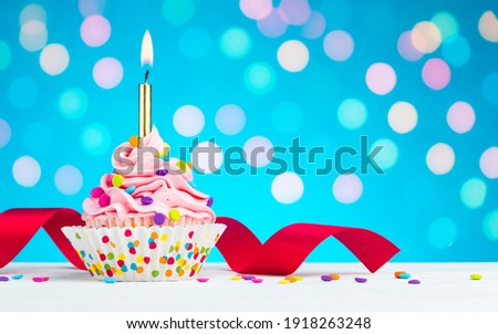 Happy Birthday cupcake. Tasty cupcakes with pink cream icing and colored sprinkles. Burning candle in a cake. Sweet delicious dessert on white wooden table and blue background with bokeh. Copy space.