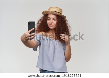 Nice looking woman, beautiful girl with curly ginger hair. Wearing striped off-shoulders blouse and hat. Taking a selfie on a smartphone. Stand isolate over white background
