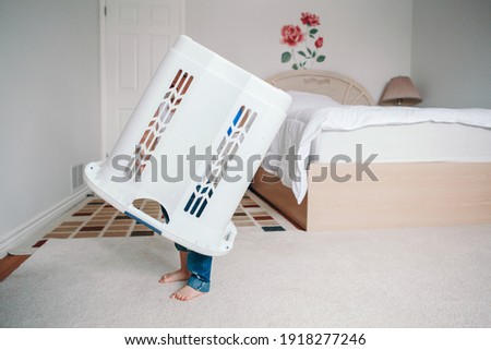 Cute funny baby toddler child with large clothes bin on head. Lonely autistic kid playing alone at home hide and seek game. Funny memorable childhood moment. Home authentic lifestyle.  Royalty-Free Stock Photo #1918277246