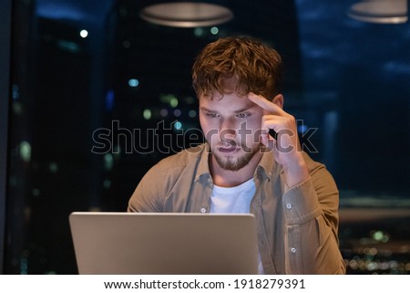 Young man analyst working hard until late hours look for rational decision for business in risky situation. Tired businessman spend evening in office by pc screen enter into important contract details Royalty-Free Stock Photo #1918279391