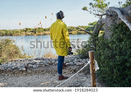 Brunette woman from back with yellow jacket observes the landscape to take pictures of the lake
