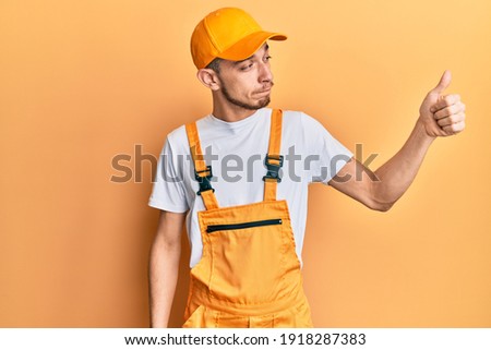 Hispanic young man wearing handyman uniform looking proud, smiling doing thumbs up gesture to the side 