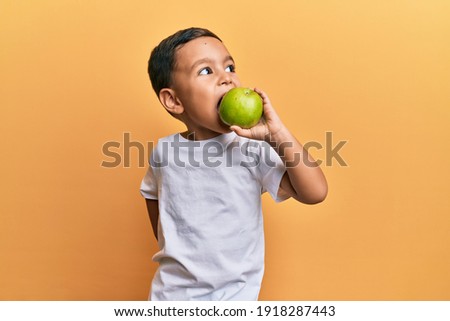 Adorable latin toddler smiling happy eating green apple looking to the side over isolated yellow background.
