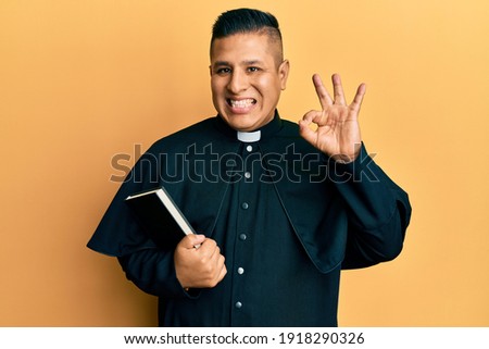 Young latin priest man holding bible doing ok sign with fingers, smiling friendly gesturing excellent symbol 
