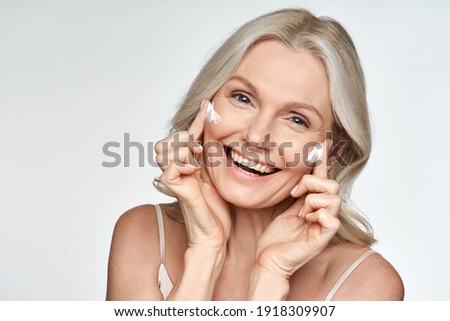 Happy 50s mid aged mature old blonde lady applying facial cream on face skin laughing enjoying anti age fresh soft skin care beauty spa treatment skincare creme isolated on white background. Portrait Royalty-Free Stock Photo #1918309907