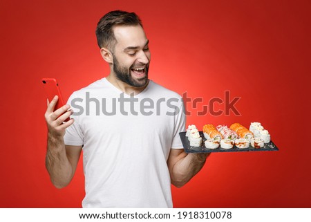 Funny young man 20s in white t-shirt using mobile cell phone typing sms message hold makizushi sushi roll served on black plate traditional japanese food isolated on red background studio portrait