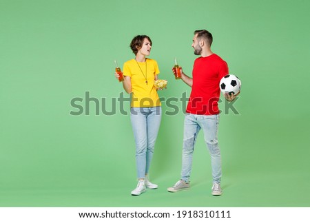 Full length displeased young couple friends sport family woman man football fans in t-shirts cheer up support favorite team with soccer ball hold beer bottles chips isolated on green background studio