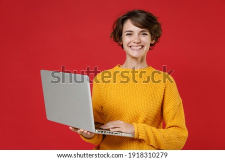Smiling cheerful beautiful young brunette woman 20s wearing basic casual yellow sweater standing working on laptop pc computer looking camera isolated on bright red colour background studio portrait