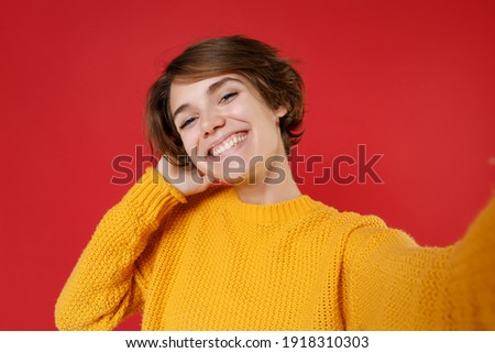 Close up of smiling attractive young brunette woman 20s in casual yellow sweater standing doing selfie shot on mobile phone put hand on head isolated on bright red colour background studio portrait
