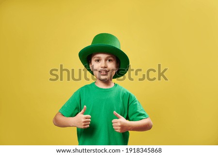 Smiling little boy in Irish green leprechaun hat showing thumbs up at camera standing on yellow background with copy space. Saint Patrick Day