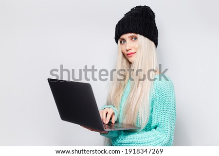 Close-up of young girl with laptop in hands against white background. Wearing black hat and blue sweater.
