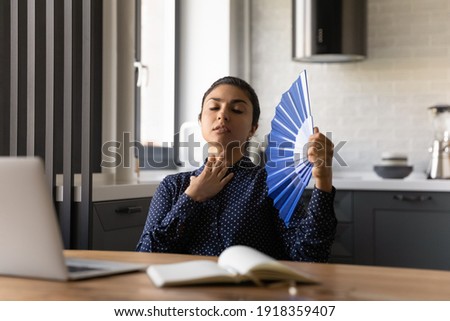 Unwell millennial Indian woman work on laptop at home office wave with hand fan feel overheated. Unhealthy tired young ethnic female use waver, suffer from hot weather or lack of AC indoors. Royalty-Free Stock Photo #1918359407