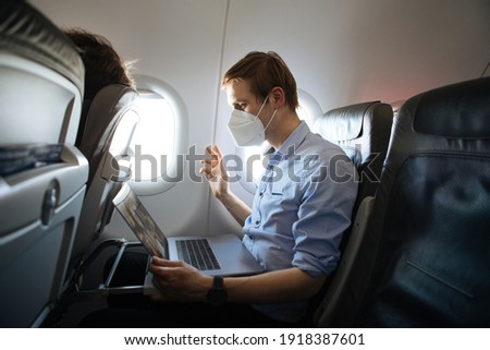 A man wearing face mask while sitting into an airplane. New normal traveling during a pandemic. Male passenger traveling. Wearing FFP2 mask in aircraft cabin. Travel Covid-19 Work from plane on laptop Royalty-Free Stock Photo #1918387601