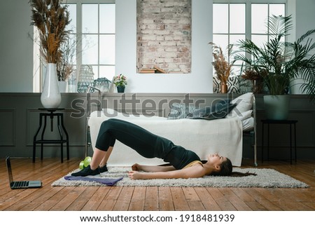 A young woman goes in for sports at home, online workout . The athlete  pumps up the buttocks, makes the gluteal bridge in the bedroom, in the background there is a bed, a vase, a carpet. Royalty-Free Stock Photo #1918481939