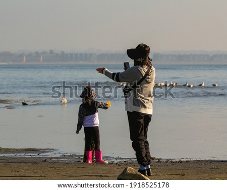 Mother and her small daughter are taking pictures with a smartphone on the beach in White Rock, BC. Travel photo, street view, selective focus.