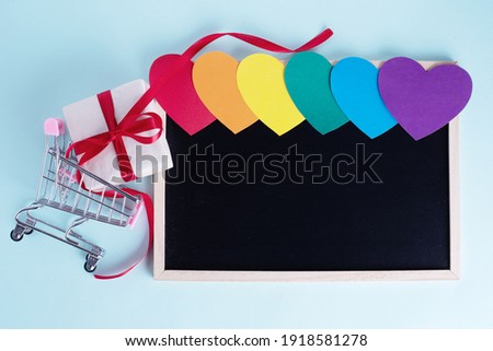 Gift box in shopping carts, multicolored rainbow colored paper hearts and a blank board with copy space on a light blue background. Seasonal sale, discounts, black friday concept. LGBT concept.
