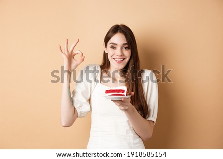 Happy birthday girl show okay gesture and hold bday cake, making wish on her holiday, standing on beige background