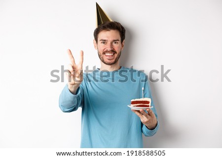 Celebration and holidays concept. Happy young man celebrating birthday, taking picture with peace sign, wearing party hat and holding bday cake, white background