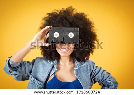 Mixed afro american woman covering eyes with retro videotape in hand with yellow background, copy space