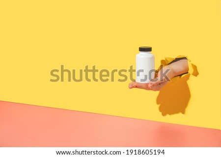 Empty white medical container for vitamins in female hand with sprinkle manicure through torn bright yellow paper background. Oral supplements, immune support, biohacking creative concept. Copy space Royalty-Free Stock Photo #1918605194