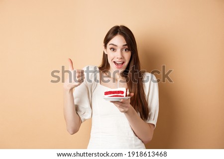 Excited birthday girl show thumb up and hold cake with candle, making wish, satisfied with bday party, smiling happy at camera, beige background