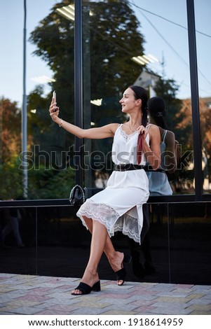 Young brunette girl with red pony tail, wearing white silk dress, sitting near modern glass building, taking selfie with phone. Pretty business woman on lunch break. Female city portrait