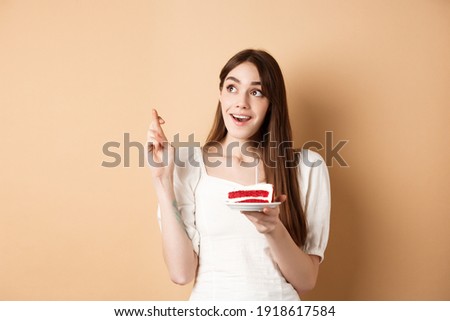 Hopeful birthday girl making wish with fingers crossed, looking up dreamy, holding bday cake with candle, beige background