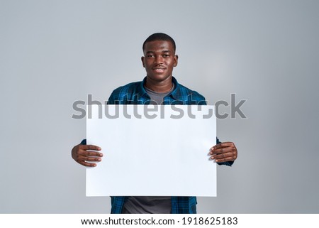 Cheerful young african american man in casual clothes smiling at camera, displaying blank banner ad, holding it in front of him, posing isolated over gray background. Front view