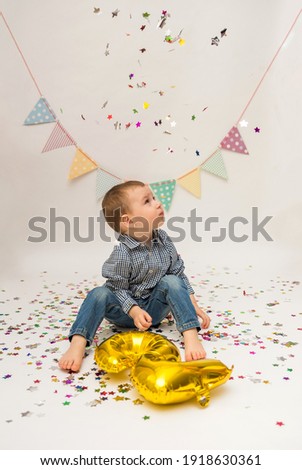 little boy is sitting in a shirt and trousers and holding a foil gold number two and looking up at the confetti on a white background