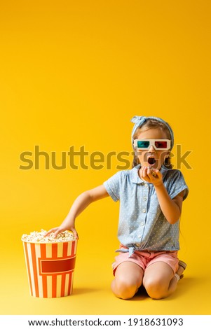amazed kid in 3d glasses eating popcorn and reaching bucket on yellow