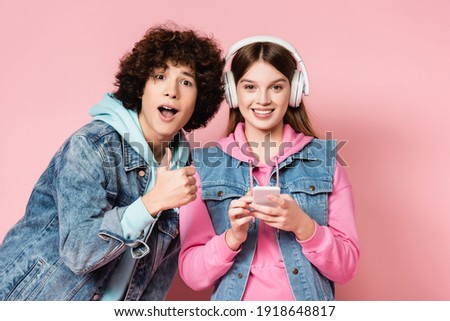 Curly teenager showing thumb up near smiling friend in headphones holding smartphone on pink background