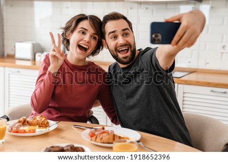 Joyful couple gesturing and taking selfie on cellphone while having breakfast at home