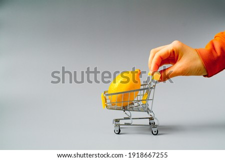 Hand with lemon in shopping cart, shopping trolley on grey background. Shopping concept. Basket of fresh lemons isolated on grey background. Vitamin C. Shopping trip.