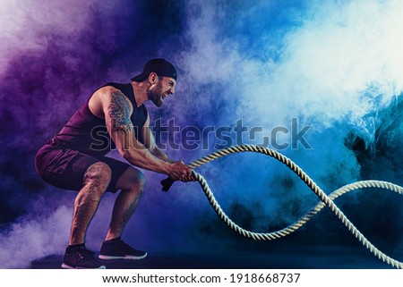 Bearded athletic looking bodybulder work out with battle rope on dark studio background with smoke. Strength and motivation. Royalty-Free Stock Photo #1918668737