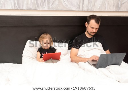 Caucasian young father is working from home with laptop in bed and caucasian little cute kid is near, watching cartoons, white linen, isolated period at lockdown with covid, remote work concept.