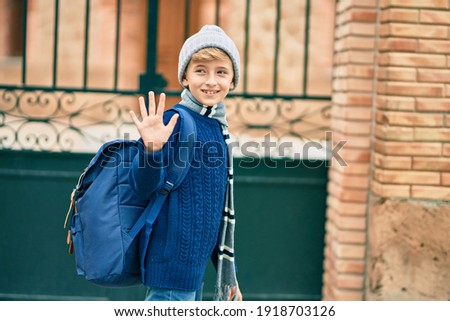 Adorable blond student kid saying goodbye with hand getting in the school.