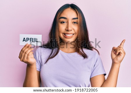 Young latin woman holding april word paper smiling happy pointing with hand and finger to the side 