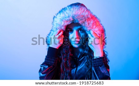 Young girl with long brown curly hair touching the hood she is wearing with her arms, against blue background