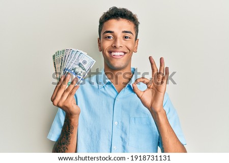 Young handsome african american man holding dollars doing ok sign with fingers, smiling friendly gesturing excellent symbol 