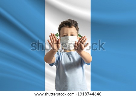 Little white boy in a protective mask on the background of the flag of Guatemala. Makes a stop sign with his hands, stay at home Guatemala.